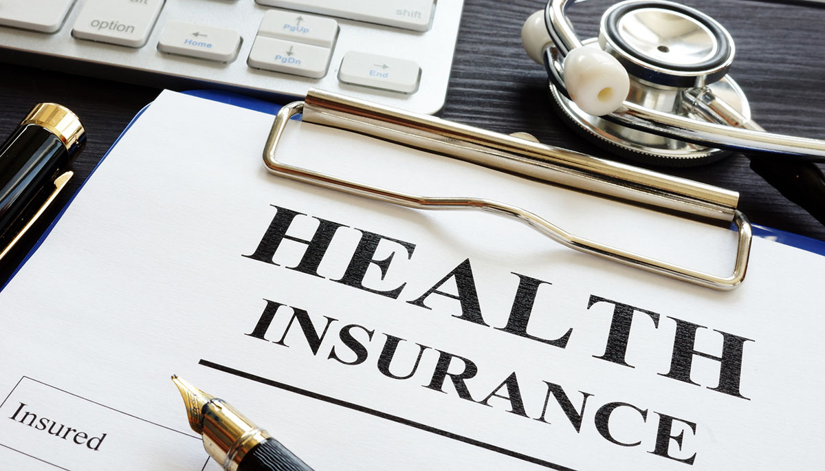 A clipboard holding a health insurance form, with a fountain pen, a stethoscope and a computer keyboard on a dark wooden desk
