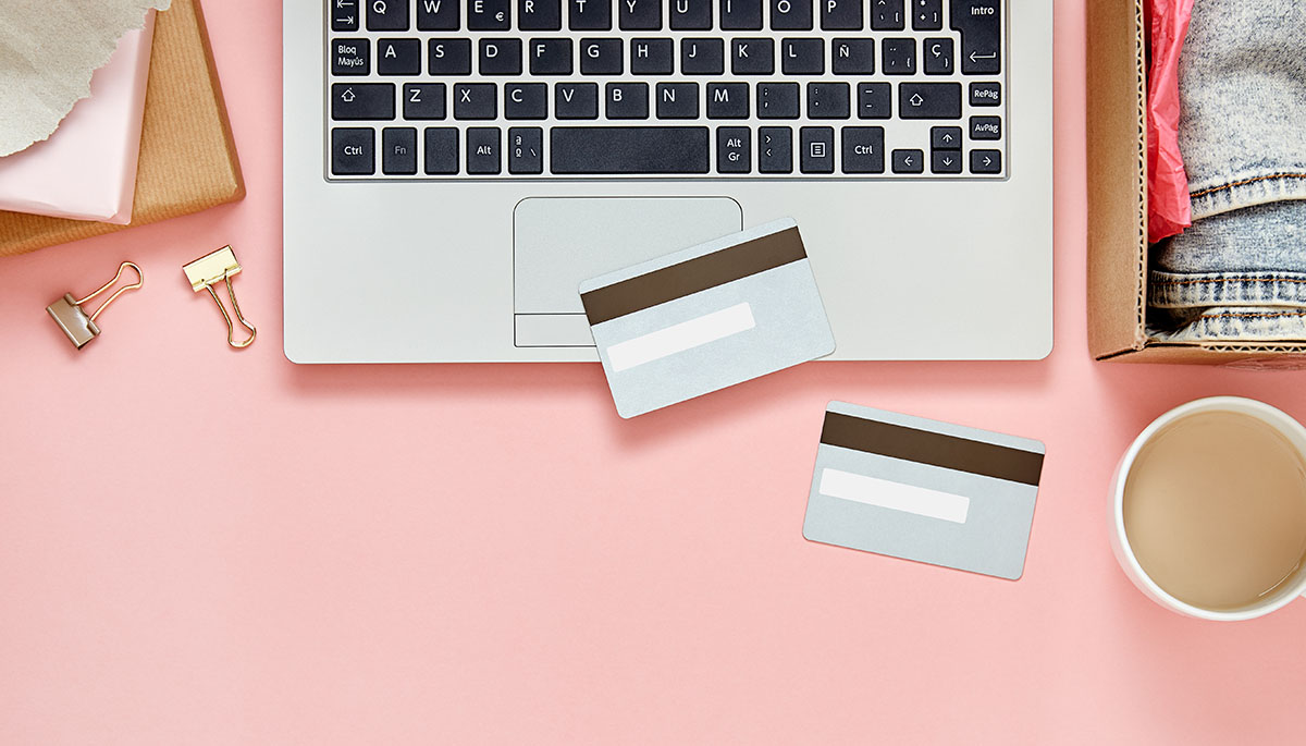 A laptop, two payment cards, a coffee cup and a parcel of folded clothes on a pink desk