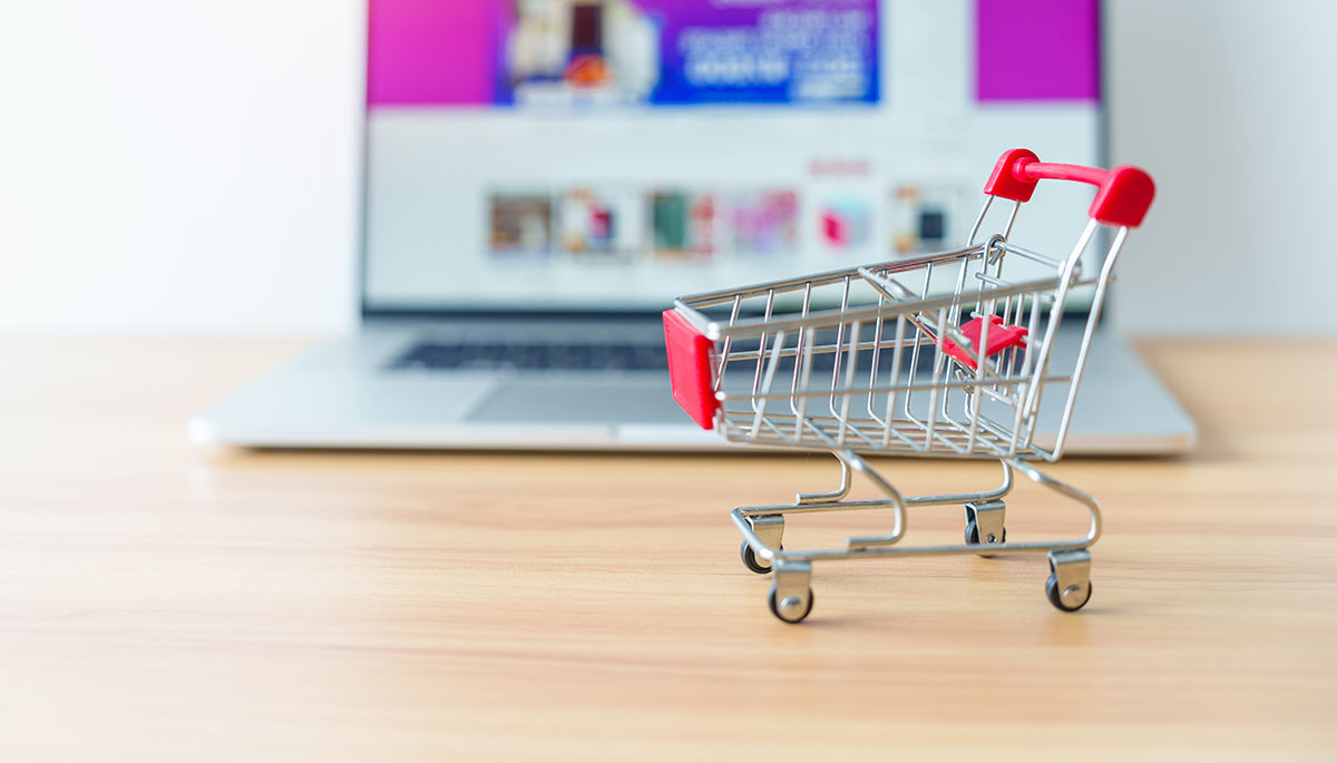 Small empty red and silver shopping cart on a wooden desk in front of a laptop showing an online store