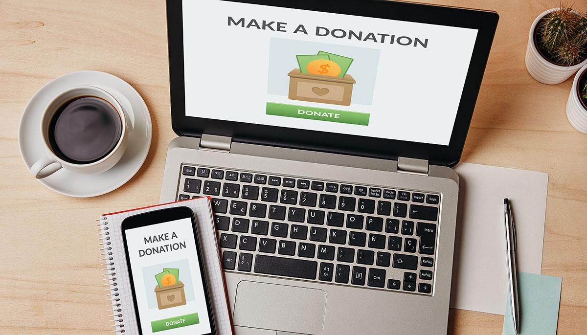 Overhead view of a laptop and a smartphone on a wooden desk, both showing a Make a Donation page with a green Donate button, beside a cup of black coffee, a notebook, a pen and a small cactus