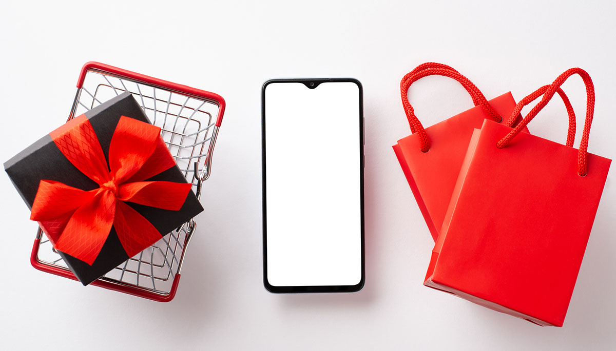 A smartphone between a shopping basket holding a wrapped gift and two red shopping bags