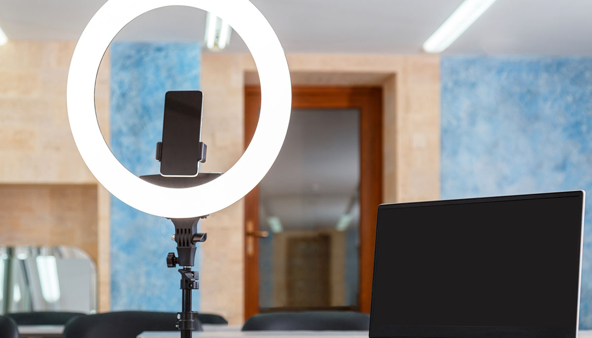 A white ring light with a smartphone mounted at its centre on a tripod, beside an open laptop