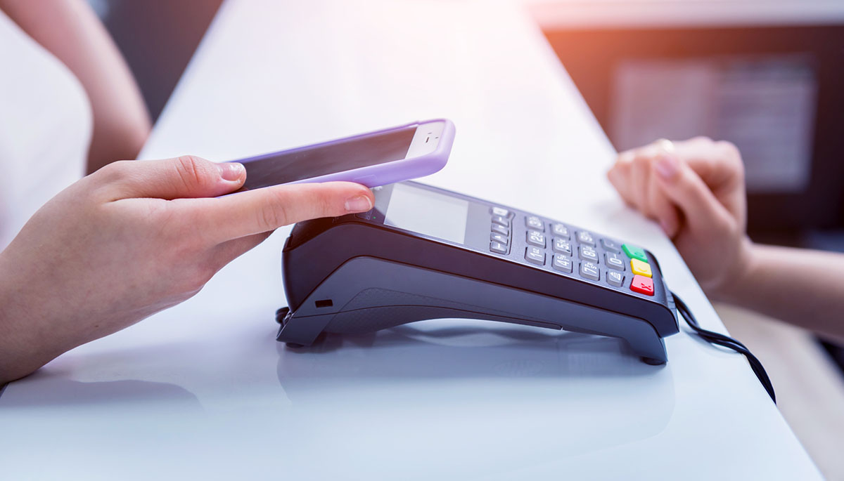 A hand holding a smartphone with a lilac case over a card payment terminal on a white counter, with another hand resting on the counter opposite